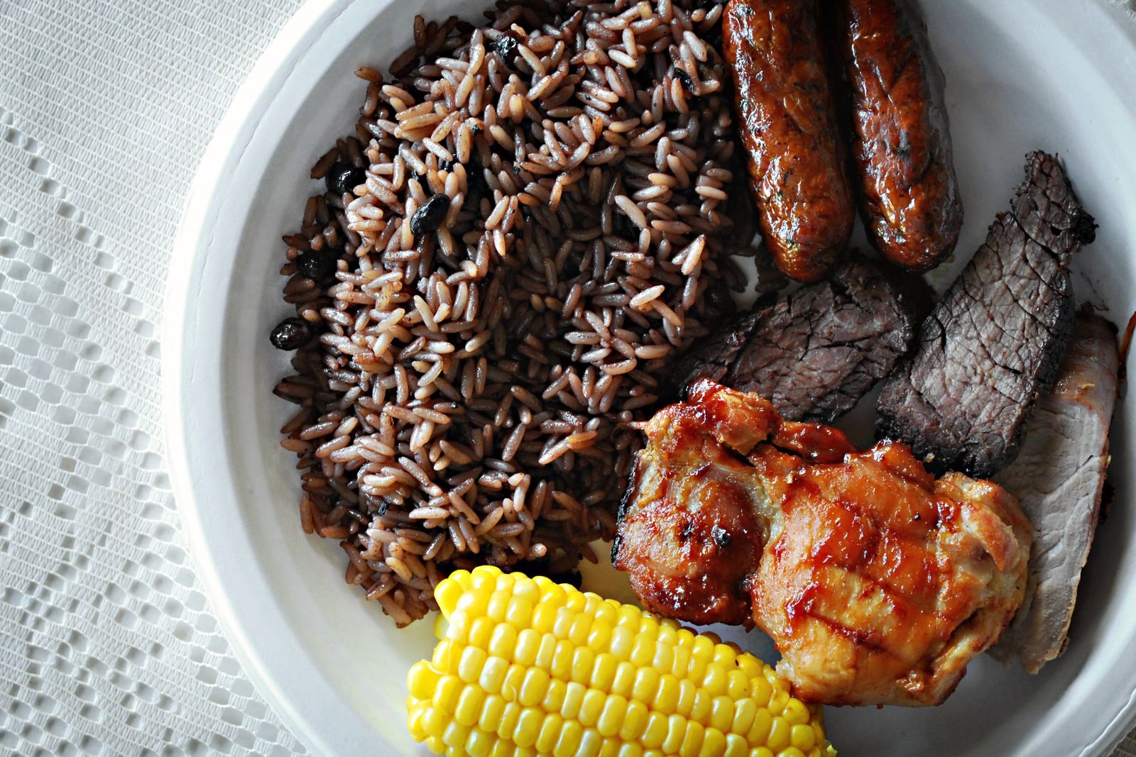 A plate featuring grilled chicken, beef, and sausages alongside seasoned rice and corn, representing a hearty meal or cultural dish.