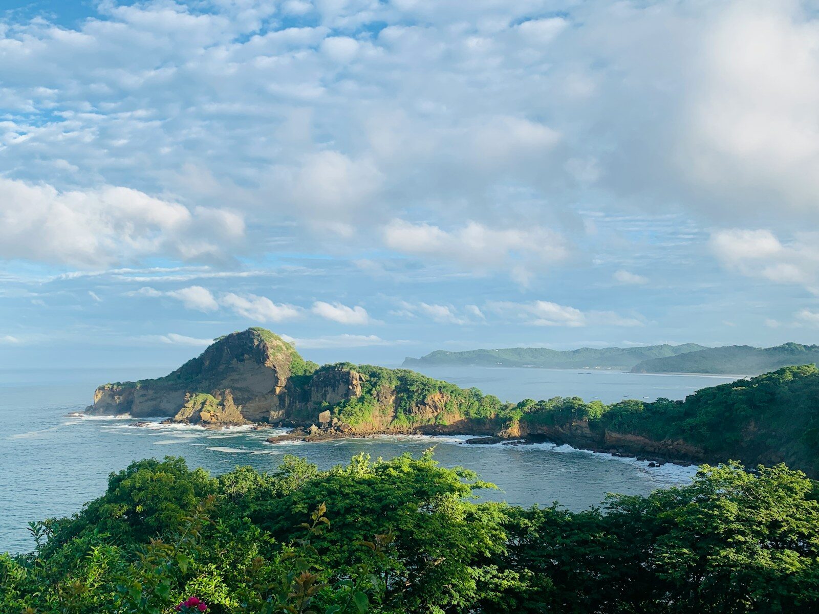 Lush coastal landscape featuring a prominent hill rising above a serene bay, with waves gently lapping against the shores under a partly cloudy sky.