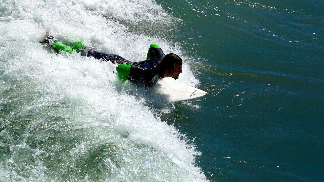 Surfer in a black wetsuit with green accents paddles on a white surfboard through turquoise waves, capturing the thrill of ocean sports.