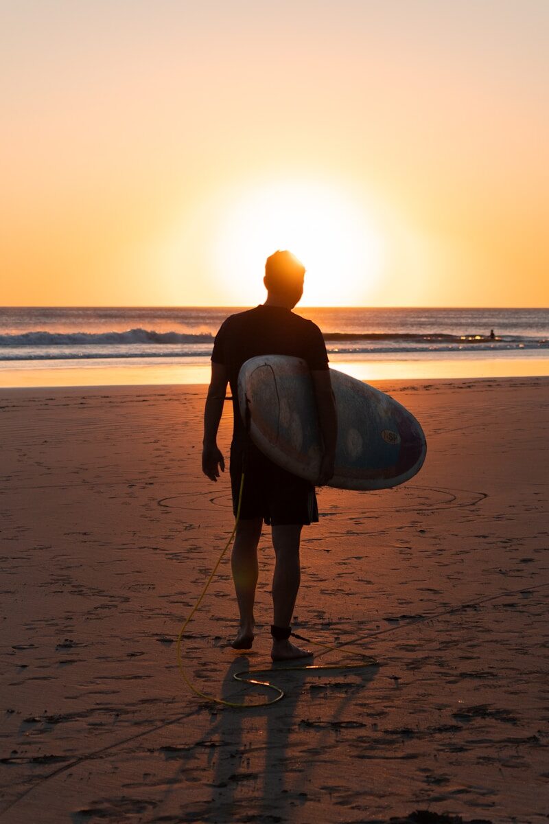 Silhouetted surfer stands on the beach at sunset, holding a surfboard, ready to enter the water amidst gentle waves.
