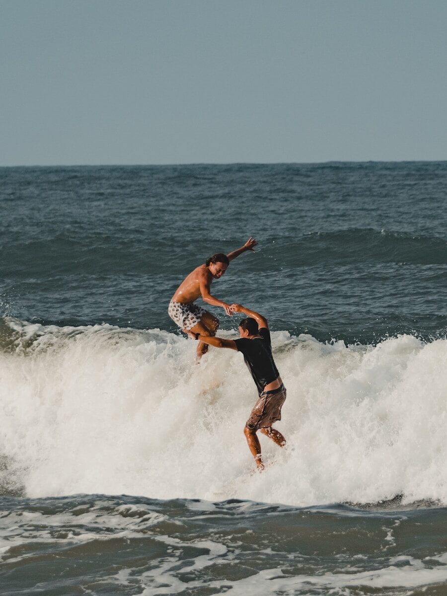 Two surfers engage in a dynamic moment as one leaps above the waves, showcasing their skill and camaraderie in the ocean surf.