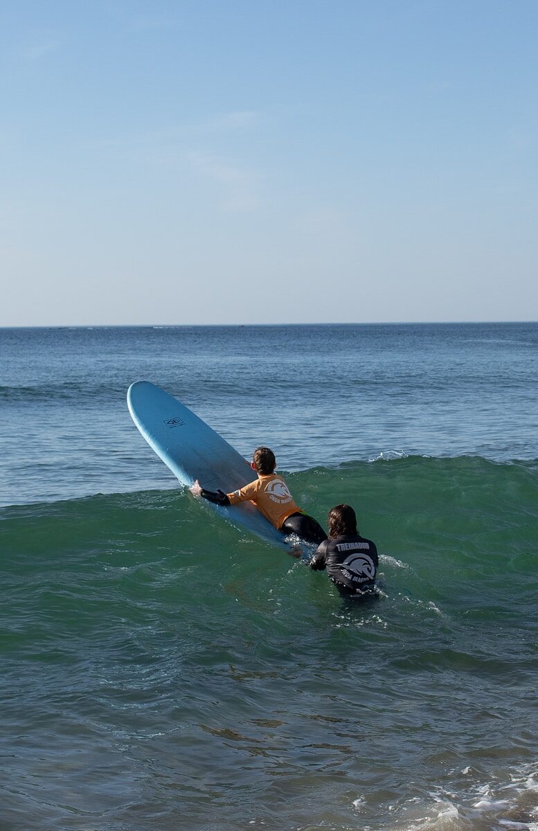 Two surfers, one in an orange rash guard and the other in black, paddle on a turquoise wave under a clear blue sky, practicing their skills.