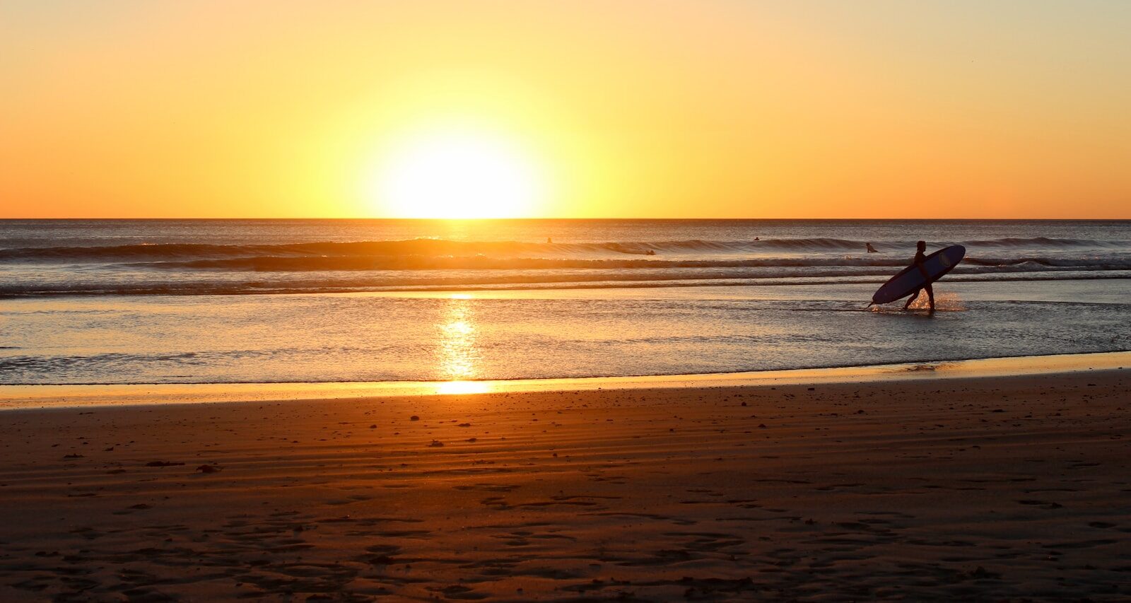 Surfer walking towards the ocean at sunset, casting a silhouette against the vibrant sky and golden waves, evoking a serene beach atmosphere.